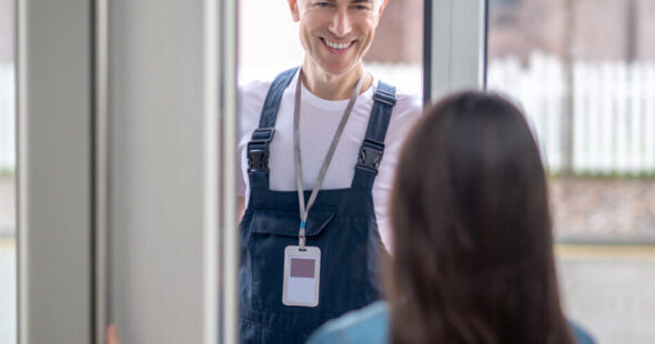 Plumber visit. Dark-haired woman with back to camera at home opening glass door to smiling middle-aged man in white tshirt and blue jumpsuit